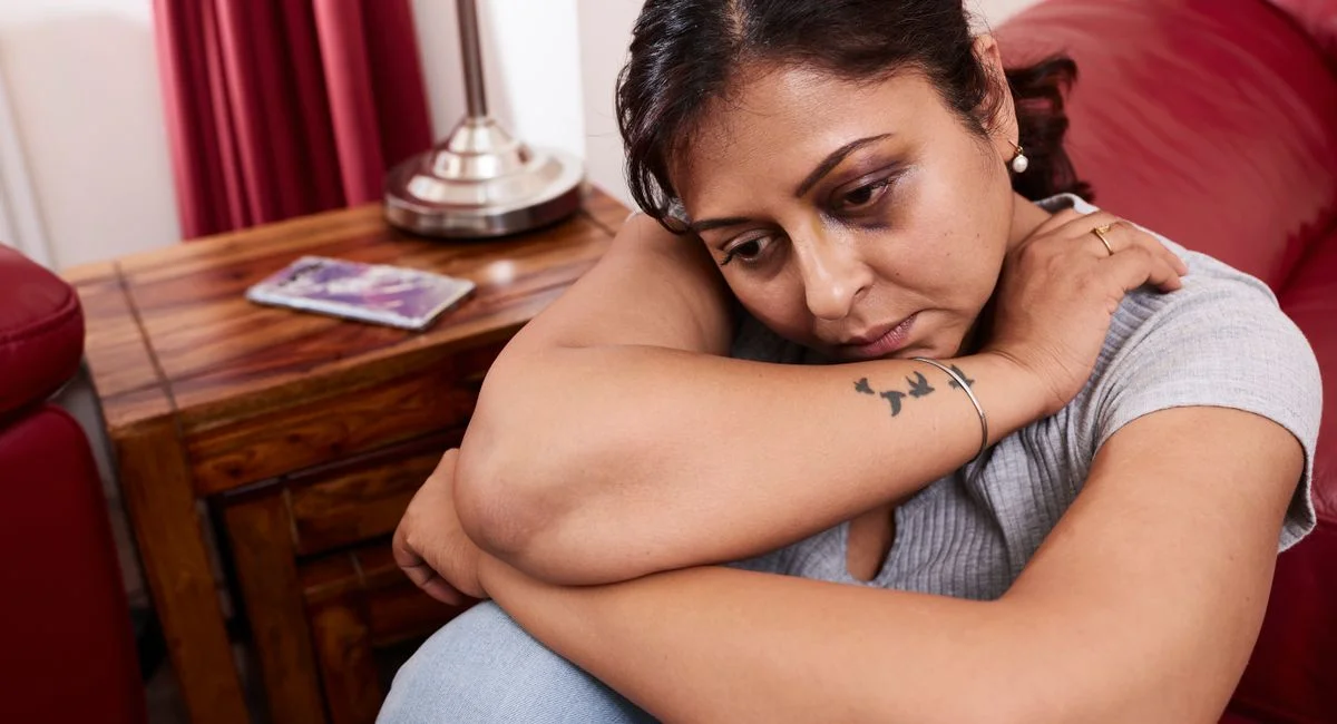 A contemplative woman with a tattoo sits indoors, reflecting on her thoughts.
