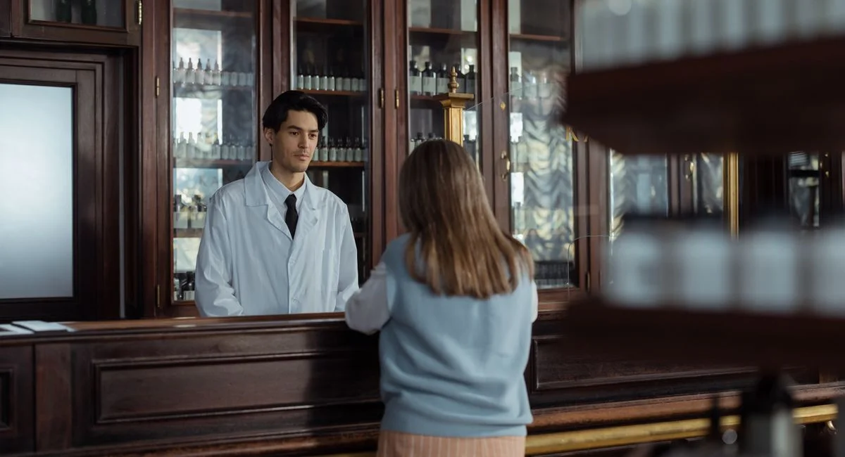 A pharmacist assists a customer at a vintage wooden pharmacy counter indoors.