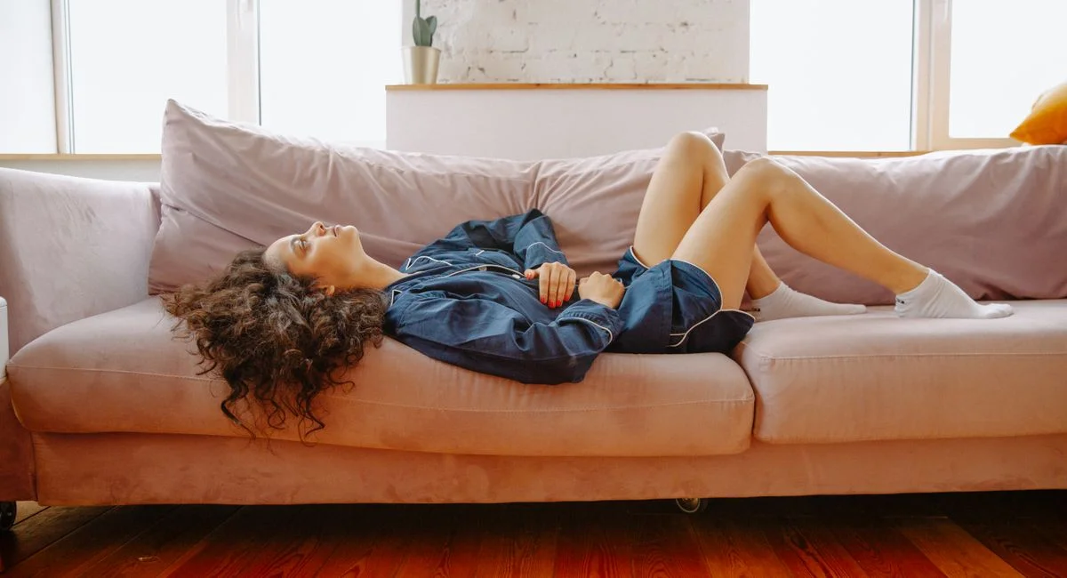 A woman in casual wear experiences abdominal discomfort while lying on a pink couch indoors.