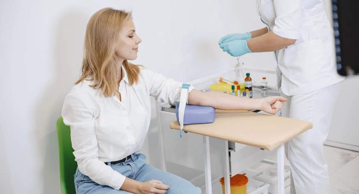 A woman sits in a clinic while a nurse prepares for a blood test procedure.