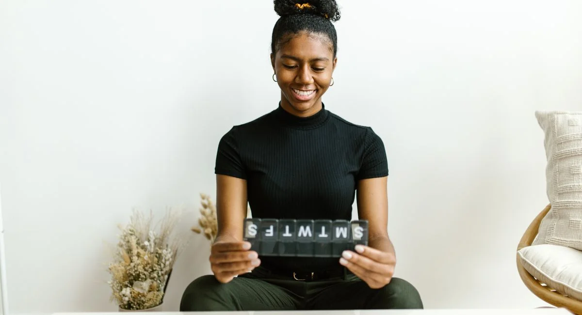 Black woman smiling while holding a weekly pill container, promoting health awareness.