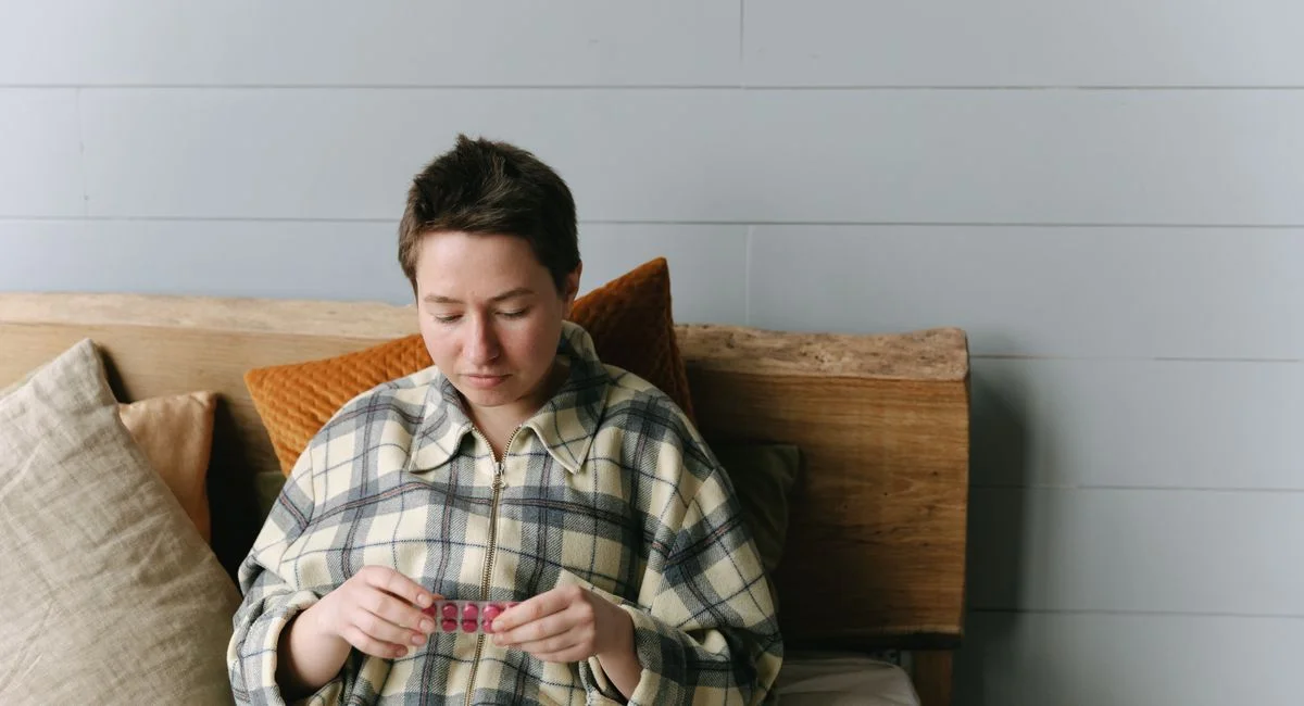 Caucasian woman in plaid shirt holds medicine blister pack while sitting comfortably indoors.
