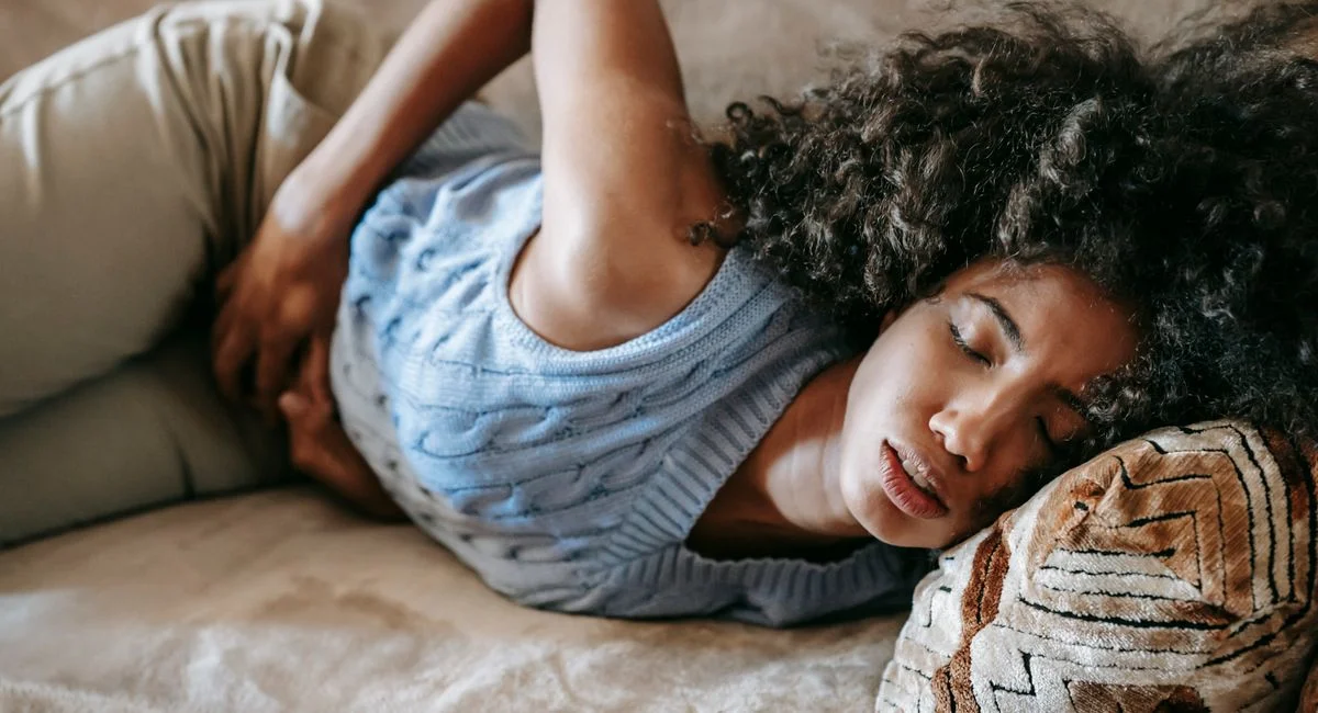 High angle of African American woman with afro hairstyle and closed eyes having pain in belly lying on sofa