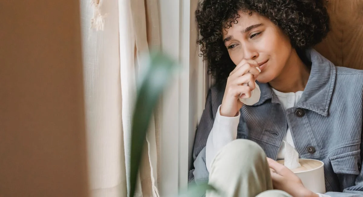 Sad African American female with tissue sitting on windowsill and crying while suffering from sorrow