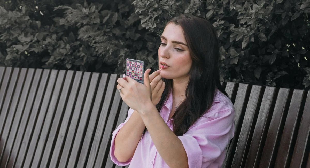 Woman in pink shirt applying lipstick with compact mirror on a bench.