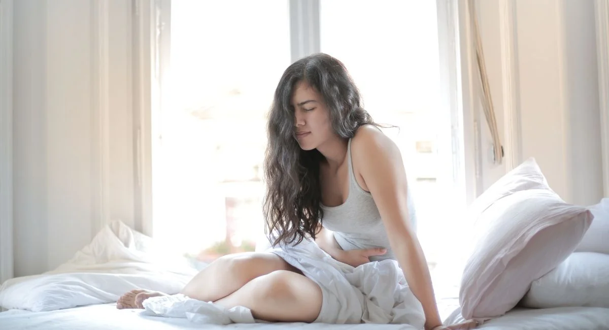 Woman sitting on bed holding her stomach, showing signs of discomfort or pain indoors.